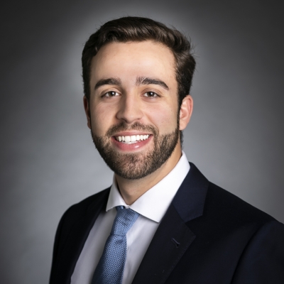 Lex Wille headshot, a white man with short dark brown hair and short facial hair. He's wearing a black suit jacket, white shirt, and blue tie. He's smiling at the camera very nicely. 